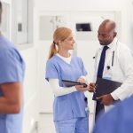 Doctor In White Coat And Nurse In Scrubs Looking At Digital Tablet In Hospital Corridor